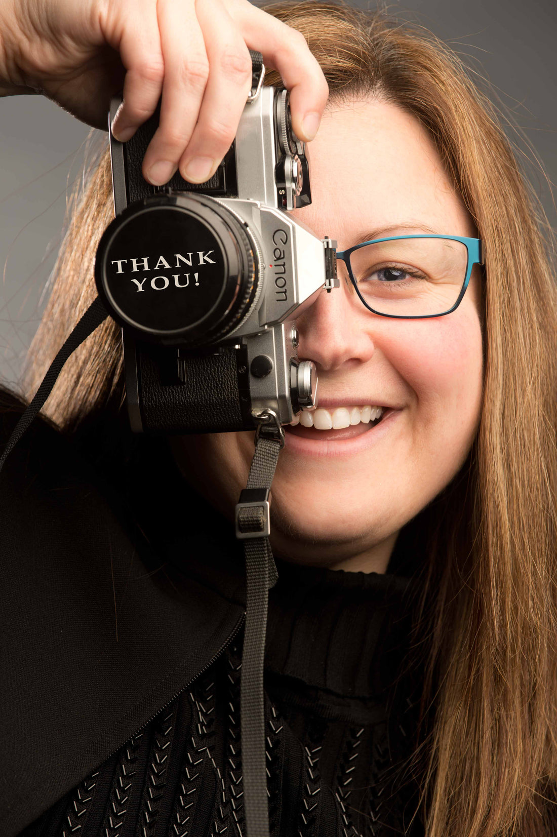 A photo of Laura Cook photographer of Vision Photography, smiling into the camera and holding up a camera and on the lens cap, Thank You is written