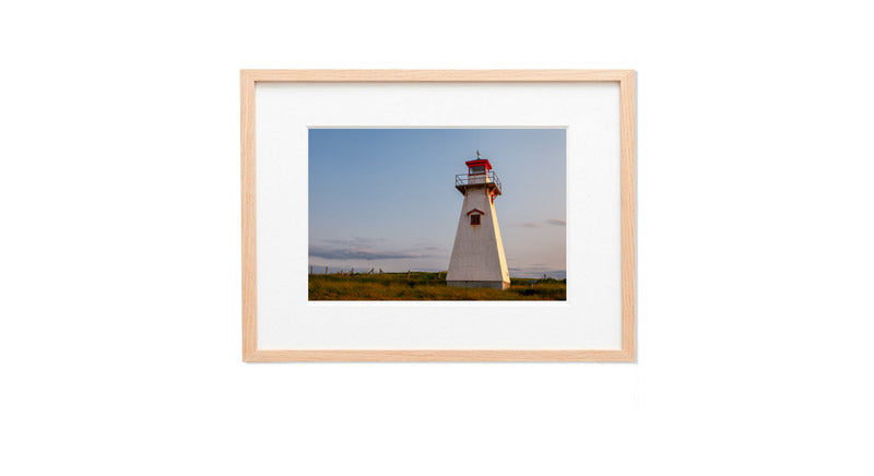 A horizontal photograph of the square, tapered white shingled Cape Tryon Lighthouse with a red lantern house, standing in a field of golden-green grass under a sunset sky.