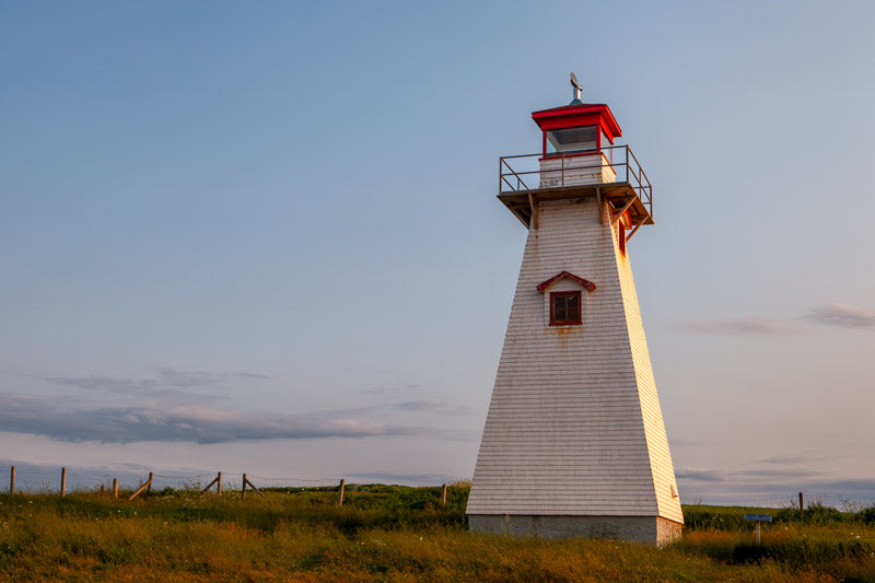 A horizontal photograph of the square, tapered white shingled Cape Tryon Lighthouse with a red lantern house, standing in a field of golden-green grass under a sunset sky.