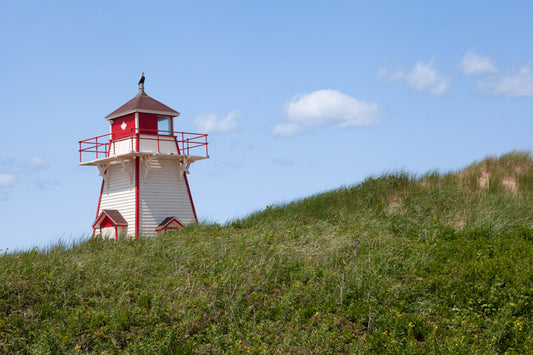 A horizontal photograph of the small, square white and red Covehead Harbour Lighthouse standing behind a grassy sand dune. The sky is bright blue with a few soft white clouds, and the foreground is filled with lush green dune grass.