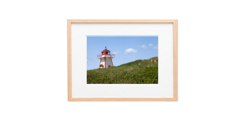 A horizontal photograph of the small, square white and red Covehead Harbour Lighthouse standing behind a grassy sand dune. The sky is bright blue with a few soft white clouds, and the foreground is filled with lush green dune grass.
