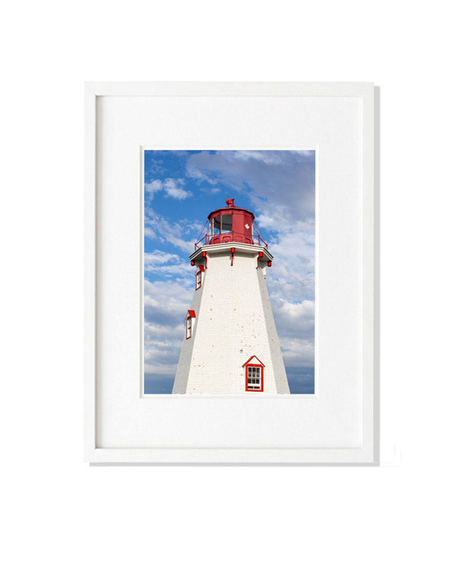 A vertical photograph of the upper portion of the white-shingled Panmure Island Lighthouse. It features a red lantern gallery with a small white maple leaf emblem, set against a bright blue sky with large, soft white and grey clouds.