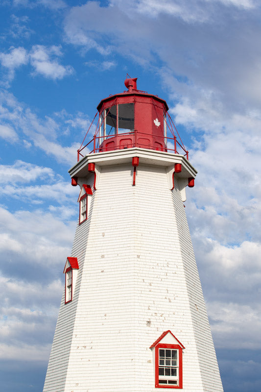 A vertical photograph of the upper portion of the white-shingled Panmure Island Lighthouse. It features a red lantern gallery with a small white maple leaf emblem, set against a bright blue sky with large, soft white and grey clouds.