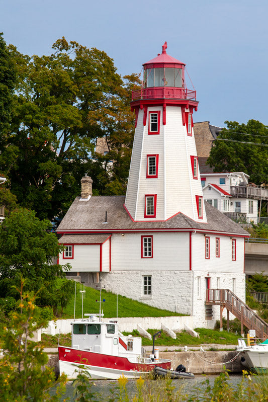 A vertical photograph of the historic white and red Kincardine Lighthouse in Ontario, featuring a matching red and white fishing boat docked in the foreground under a clear blue sky.