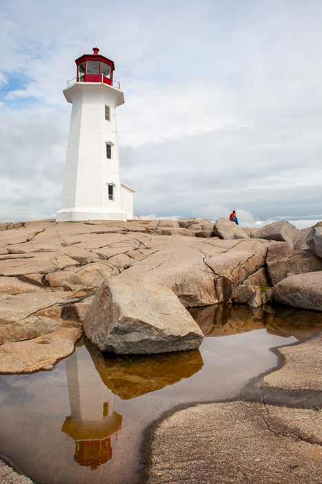 eggy’s Cove Lighthouse on rugged granite rocks during the day, with its perfect reflection captured in a calm tide pool. A single figure sits in the background under a partly cloudy sky.