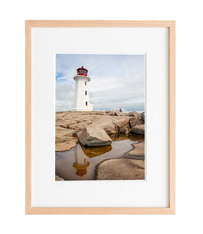 eggy’s Cove Lighthouse on rugged granite rocks during the day, with its perfect reflection captured in a calm tide pool. A single figure sits in the background under a partly cloudy sky.