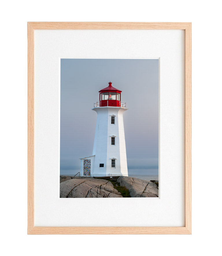 A vertical photograph of Peggy’s Point Lighthouse at twilight. The white octagonal tower stands on smooth, grey granite rocks against a soft, hazy grey-blue sky. The red lantern at the top is vibrant, and the small door at the base is covered in colorful stickers.