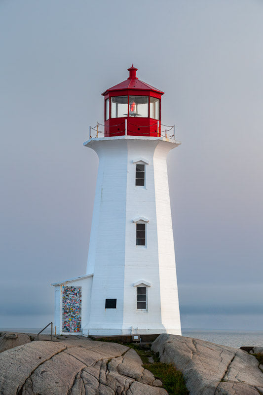 A vertical photograph of Peggy’s Point Lighthouse at twilight. The white octagonal tower stands on smooth, grey granite rocks against a soft, hazy grey-blue sky. The red lantern at the top is vibrant, and the small door at the base is covered in colorful stickers.