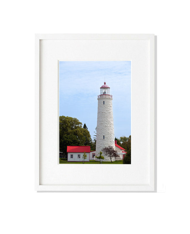 A vertical photograph of the tall, white limestone Point Clark Lighthouse with a red lantern. Beside the tower is a small white building with a vibrant red metal roof, surrounded by green trees under a soft, overcast sky.