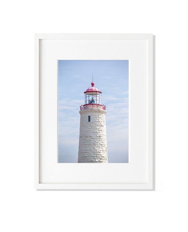 A vertical close-up photograph of the upper section of the Point Clark Lighthouse, focusing on the lantern room. The tower is constructed of off-white textured limestone blocks, topped with a red lantern gallery and a pinkish-red dome against a pale blue sky.