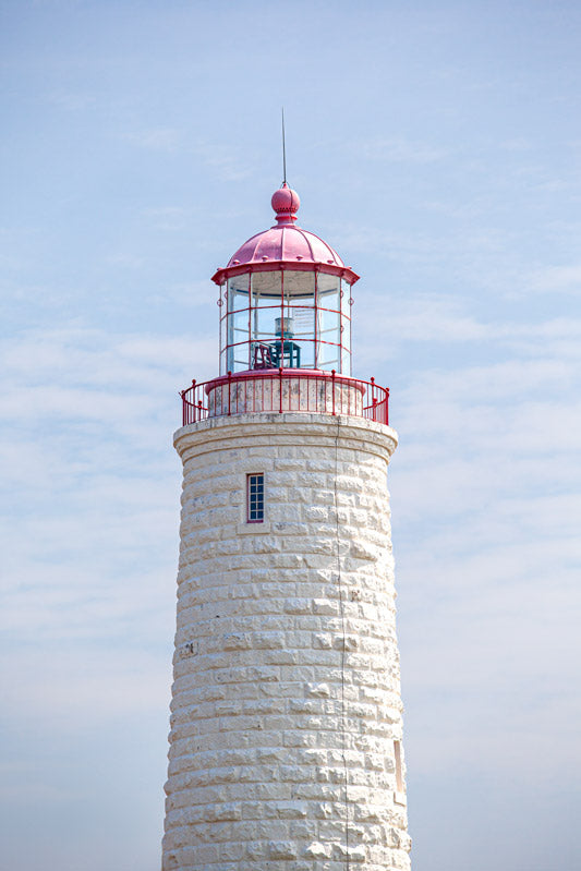 A vertical close-up photograph of the upper section of the Point Clark Lighthouse, focusing on the lantern room. The tower is constructed of off-white textured limestone blocks, topped with a red lantern gallery and a pinkish-red dome against a pale blue sky.