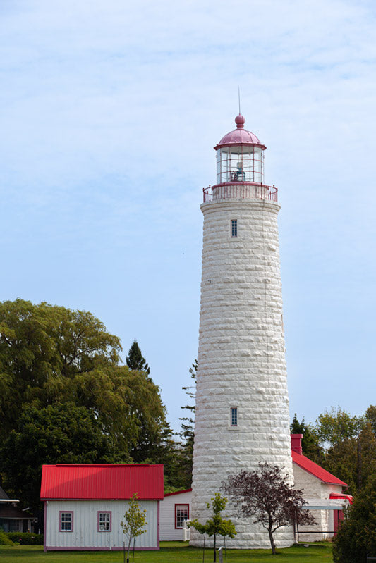 A vertical photograph of the tall, white limestone Point Clark Lighthouse with a red lantern. Beside the tower is a small white building with a vibrant red metal roof, surrounded by green trees under a soft, overcast sky.