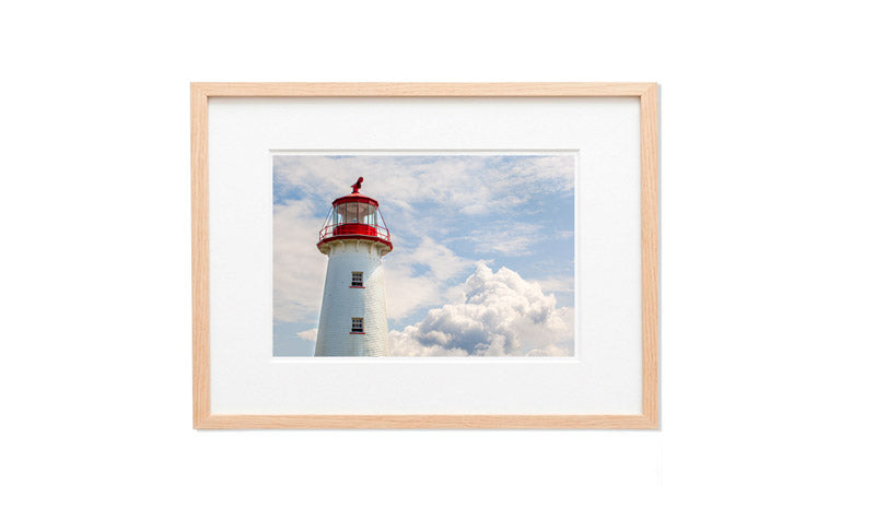 A horizontal photograph focusing on the upper half of the white shingled Point Prim Lighthouse with its red lantern gallery. A large, fluffy white cloud sits to the right of the tower against a pale blue sky with light, wispy clouds.