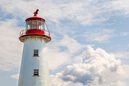 A horizontal photograph focusing on the upper half of the white shingled Point Prim Lighthouse with its red lantern gallery. A large, fluffy white cloud sits to the right of the tower against a pale blue sky with light, wispy clouds.