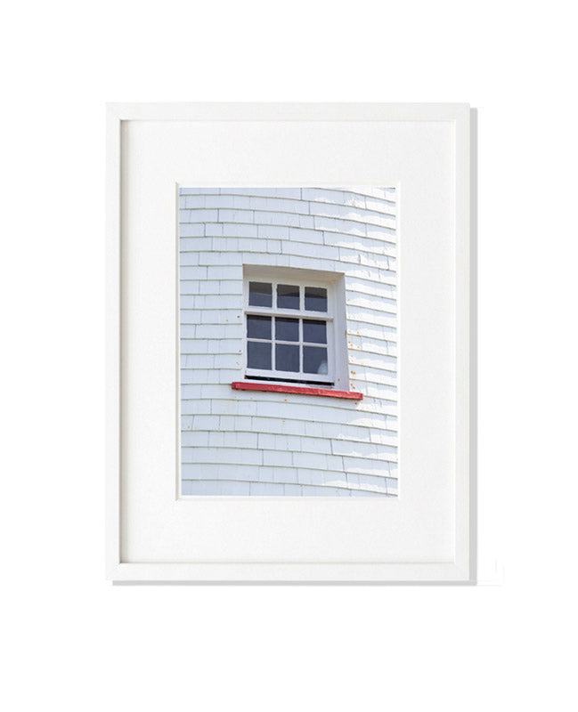 A vertical close-up photograph of a white-framed window set into the white shingled exterior of Point Prim Lighthouse. A thin red windowsill sits at the bottom, and the shingles show weathered texture and slight aging.