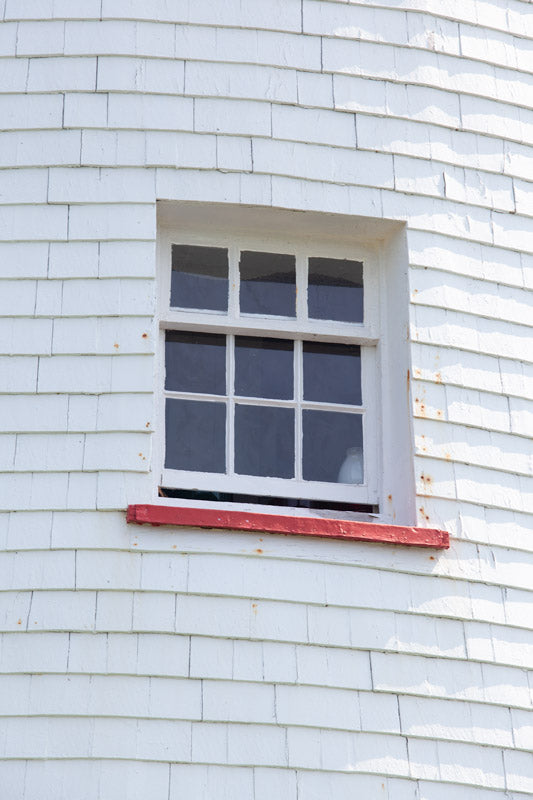 A vertical close-up photograph of a white-framed window set into the white shingled exterior of Point Prim Lighthouse. A thin red windowsill sits at the bottom, and the shingles show weathered texture and slight aging.