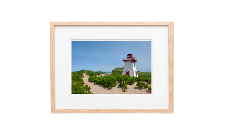 A horizontal framed photograph of the white and red St. Peter’s Harbour Lighthouse on PEI, surrounded by green beach grass and a sandy path under a blue sky.