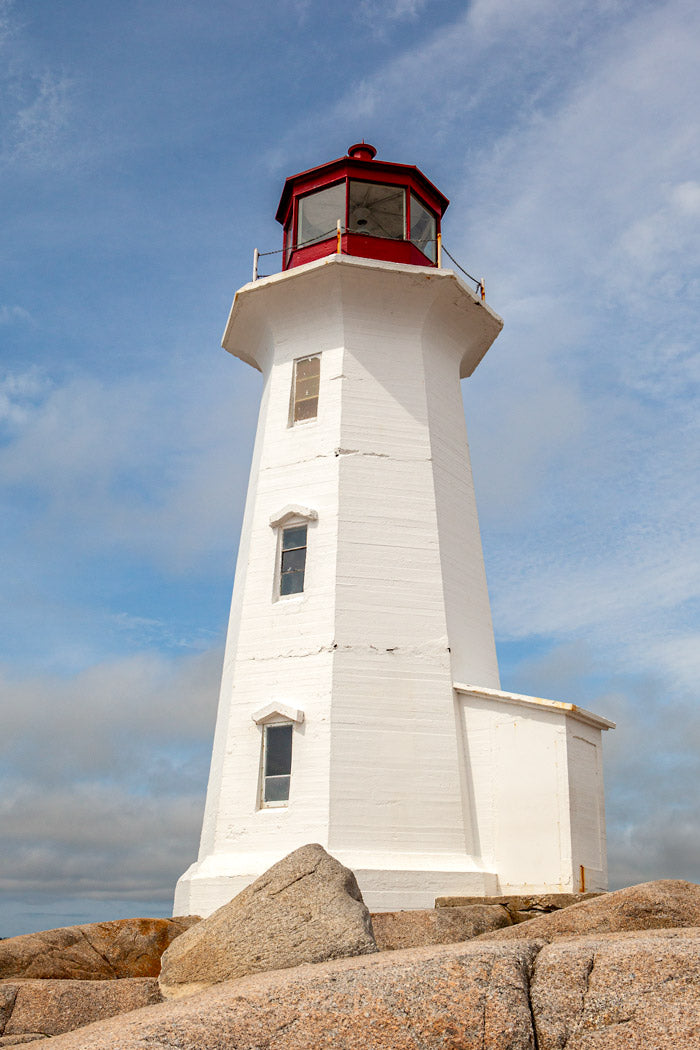 Peggy's Cove Lighthouse with a red lantern house, standing on large granite rocks under a bright blue sky with soft white clouds.