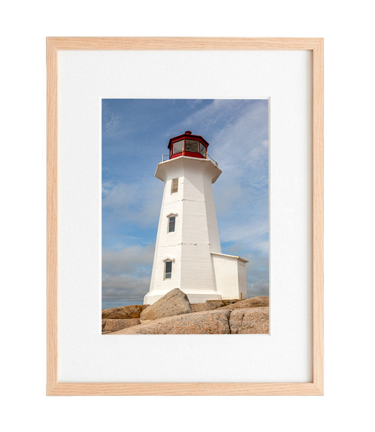 Peggy's Cove Lighthouse with a red lantern house, standing on large granite rocks under a bright blue sky with soft white clouds.