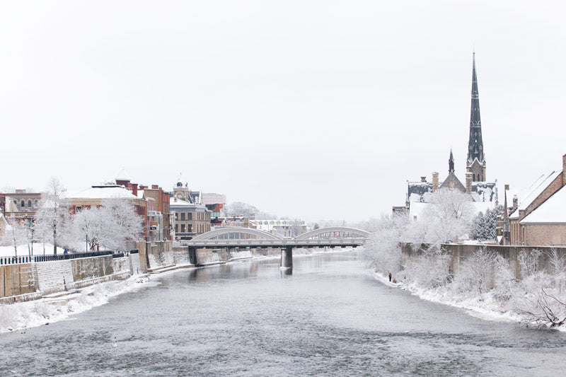 A peaceful winter landscape of the Grand River in Galt, Cambridge, featuring snow-covered trees, historic buildings, and a church spire under a soft white sky.