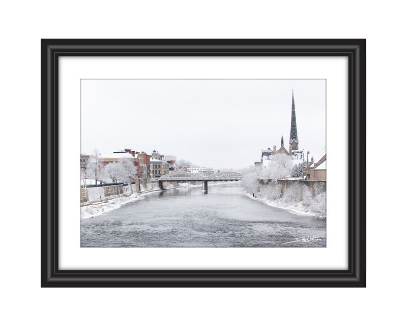 A peaceful winter landscape of the Grand River in Galt, Cambridge, featuring snow-covered trees, historic buildings, and a church spire under a soft white sky.