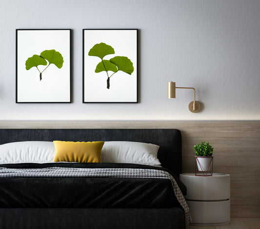 a picture of a bedroom wiht a bed and above the bed on the wall is 2 frames with images of green ginko leaves on white backgrounds
