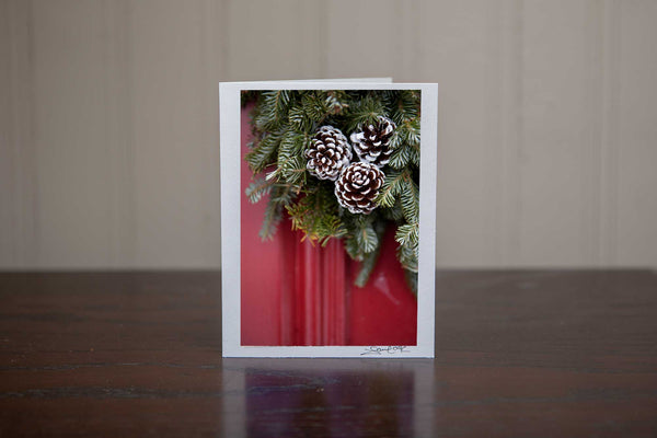 Greeting card featuring 'Adorn' a photograph of a red door with close up of a Christmas wreath of evergreen trees and pinecones on the door Photo by Cambridge Ontario Photographer Laura Cook of Vision Photography